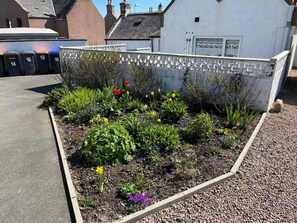 Un parterre fleuri avec différentes plantes et fleurs, un mur blanc orné d’un motif décoratif et, au fond, un bâtiment avec un garage.
