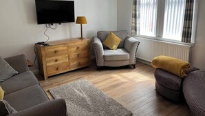 A living room with a flat-screen TV, wooden dresser, grey sofa, armchair, and a window with curtains. - Costal Cottage -boddam-sleeps6- Pet Friendly (Peterhead)