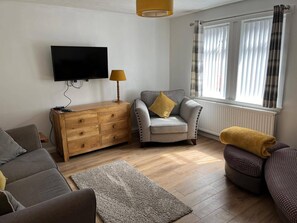 A living room with a flat-screen TV, wooden cabinet, grey sofa, armchair with a yellow pillow, and a window with curtains. - Costal Cottage -boddam-sleeps6- Pet Friendly (Peterhead)