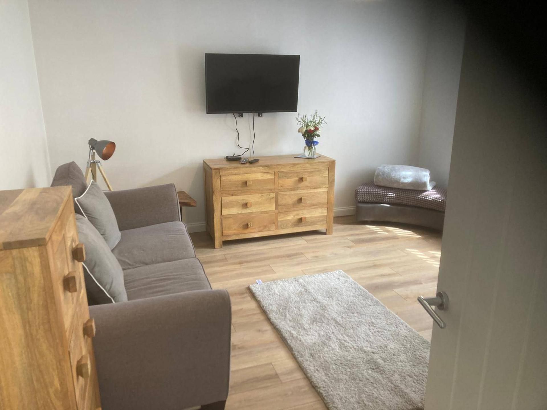 Uma sala de estar com um aparador de madeira, um sofá cinza, uma TV de tela plana fixada na parede e uma pequena mesa com um vaso de flores.