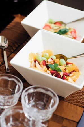 Two white bowls of fruit salad with a spoon, placed on a wooden table with a spoon and two glasses. - Olivers Bed and Breakfast (Mtunzini)