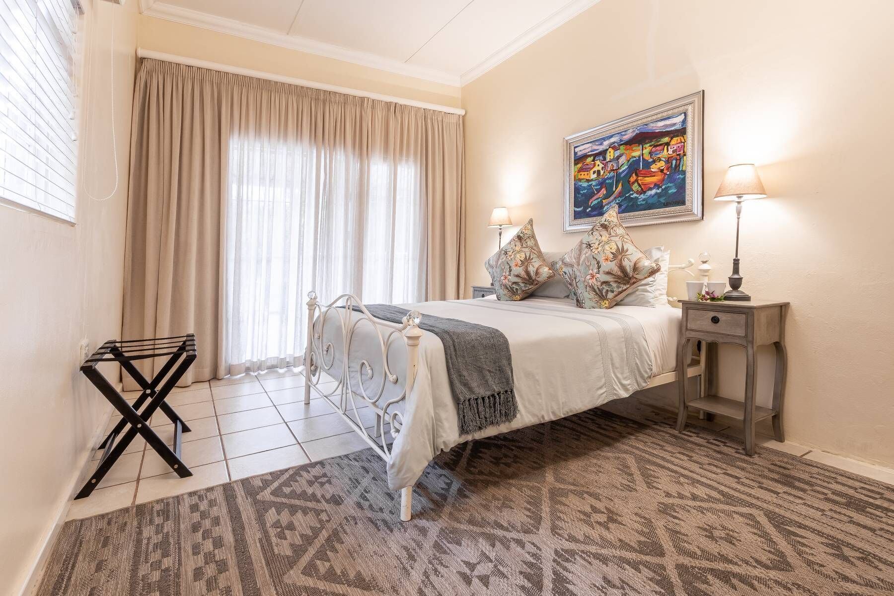 A hotel room with a bed, a nightstand, a lamp, a painting, and a window with curtains.