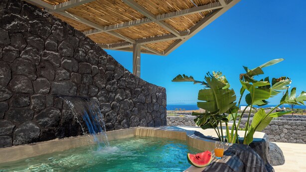 A hot tub with a waterfall feature, surrounded by tropical plants and a stone wall, overlooking a clear blue sky and ocean. - Mperes Villas (Santorini)