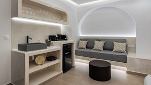 A modern kitchen with a built-in sink, a coffee machine, and a small refrigerator. There is a gray sofa with patterned cushions against the wall, and a round black ottoman in the center. - Mperes Villas (Santorini)
