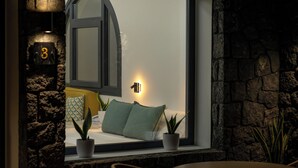 A cozy room with a large window, a stone wall, a bed with pillows, and a small potted plant. - Mperes Villas (Santorini)
