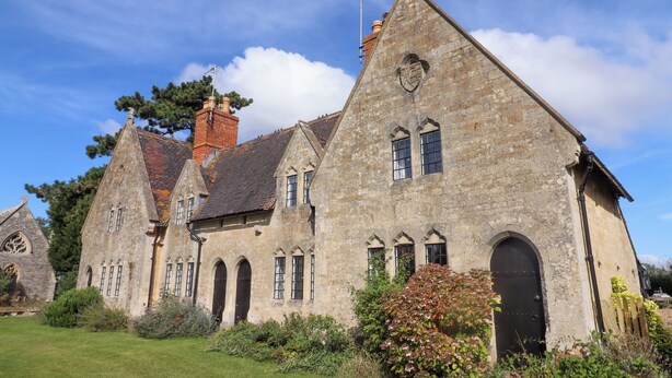 A stone building with multiple arched windows and a central door, surrounded by a well-maintained lawn and some shrubs. - The Alms House East (Gloucester)