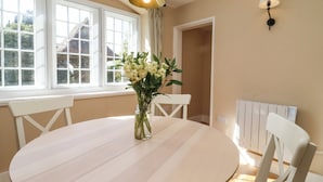 A dining area with a round table, white chairs, a vase with flowers, a hanging light fixture, and a radiator. - The Alms House East (Gloucester)