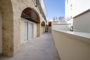 Un couloir étroit, carrelé, avec des portes en arc donnant sur un bâtiment à la façade blanche et doté d’un balcon.