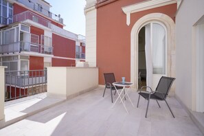 A balcony with a table and two chairs, a red building in the background, and a white door.