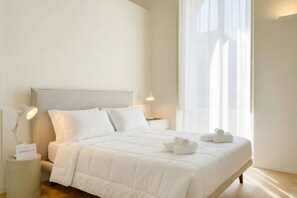 Ein ordentlich bezogenes Bett mit weißen Laken, zwei Nachttischlampen und ein Fenster mit durchscheinenden Vorhängen.
