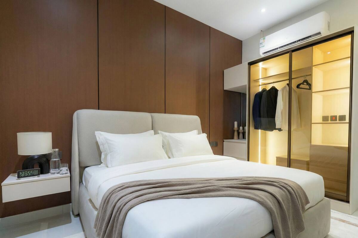 A modern hotel room with a large bed, a nightstand with a lamp, a wardrobe with a mirror, and a coat hanging inside.