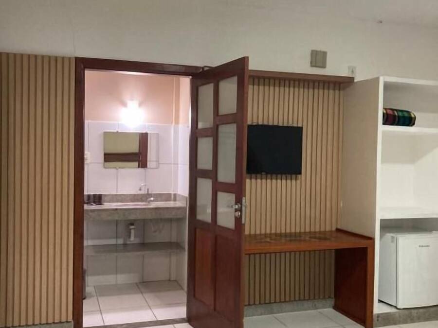 A room with a tiled floor, a wooden door leading to a bathroom, a white shelf unit, and a flat-screen TV mounted on the wall.