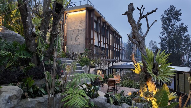 A modern building with a stone facade, surrounded by lush greenery and a wooden deck with outdoor seating.