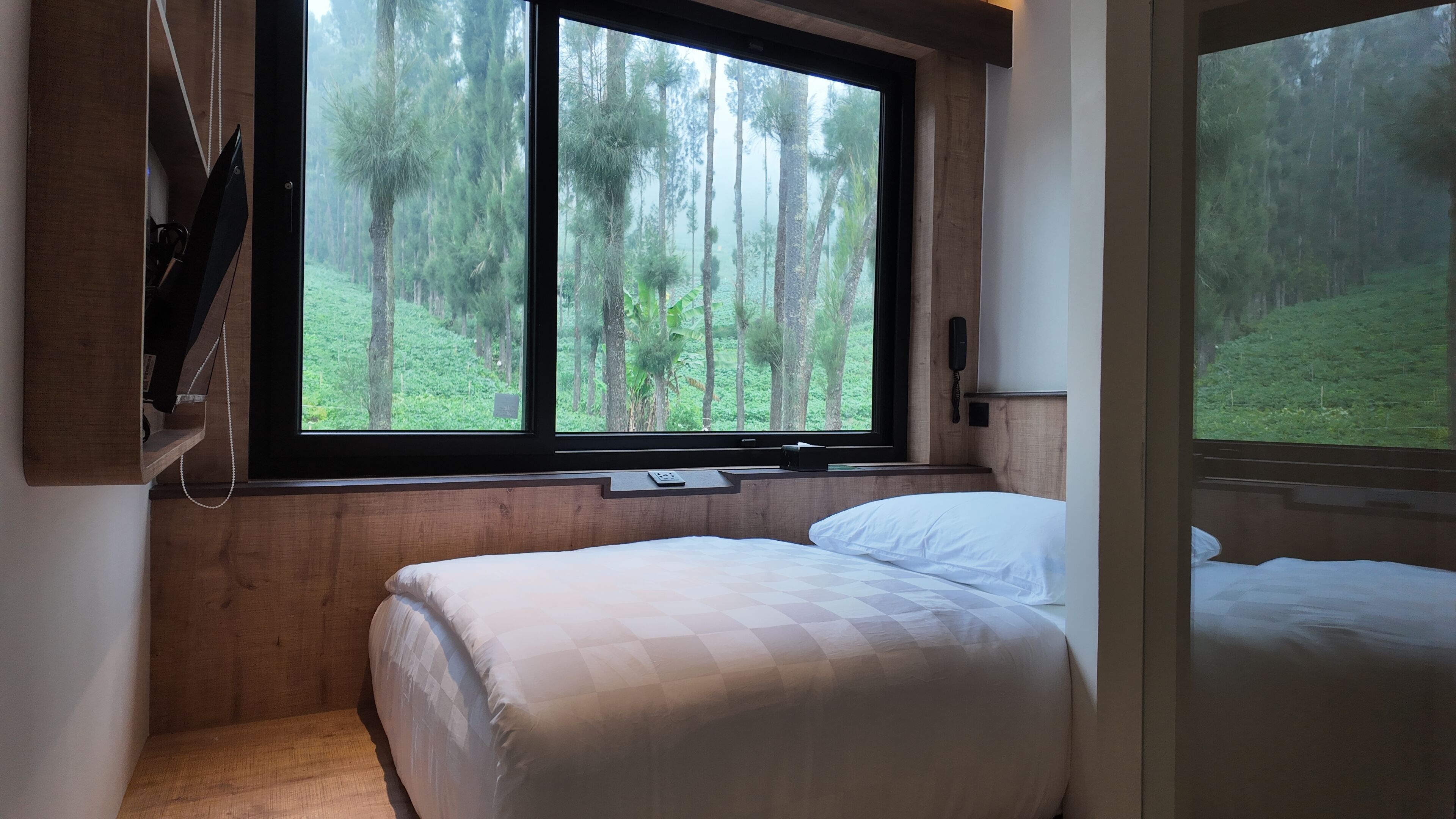 A bedroom with two beds, a large window with a forest view, a bedside table, and a phone.