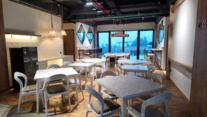 A modern dining area with white tables and chairs, a black refrigerator, and large windows offering a view of the outdoors. - Jambuluwuk Bromo Poshtel (Wonokitri)