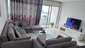 A living room with a grey sectional sofa, a glass coffee table, and a flat-screen TV on a white stand. - Sublime T2 Le Plateau Lagoon view Bright Large Ventilated (Abidjan)