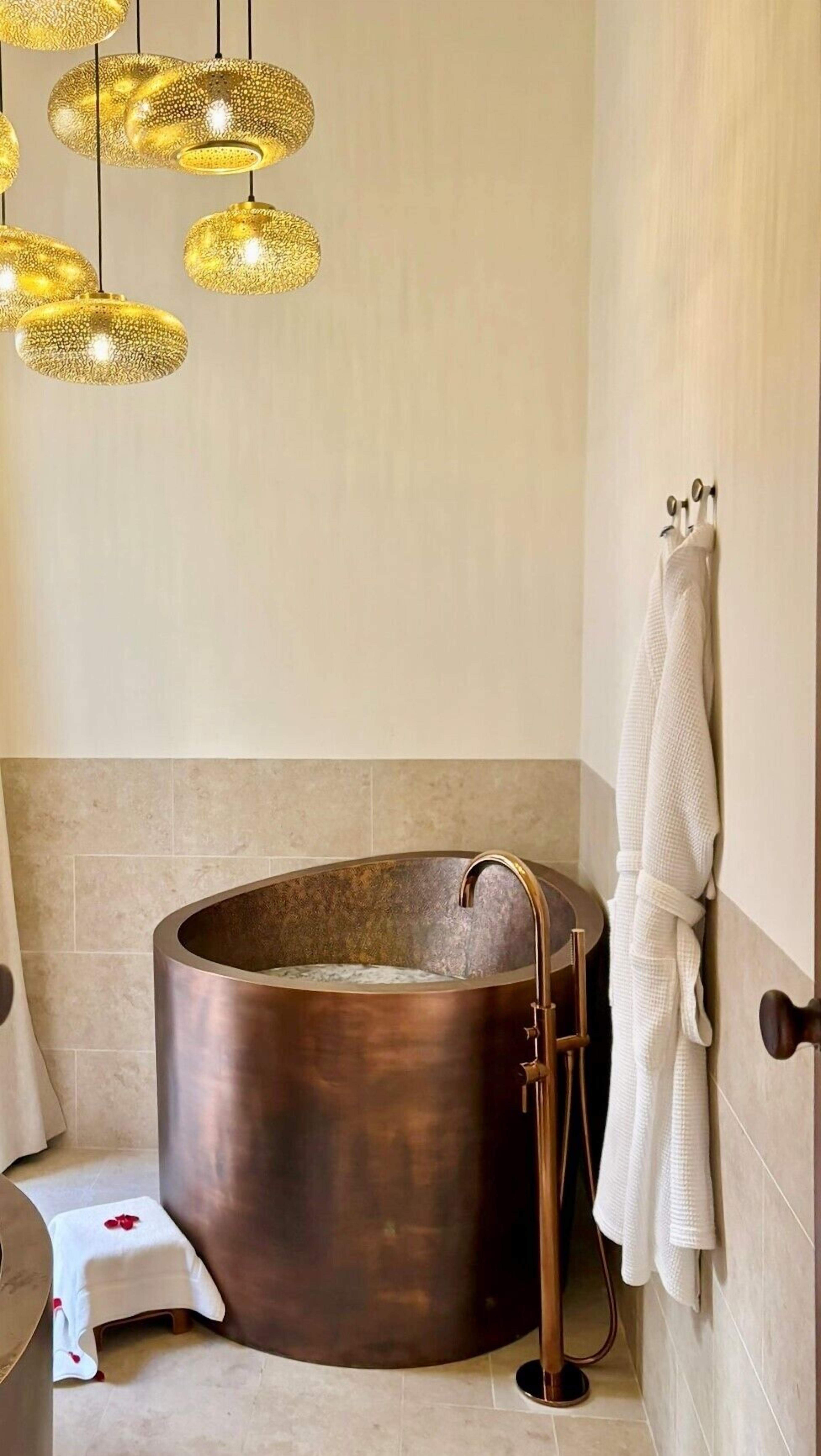 Een moderne badkamer met een groot, rond koperbad, een witte handdoekhouder en een bijzondere lichtarmatuur met een structuur.