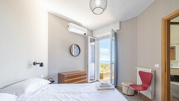 A bedroom with a bed, a dresser, a red chair, a radiator, and a balcony with a view. - Sun & Sea Homes by Wonderful Italy (Sanremo)
