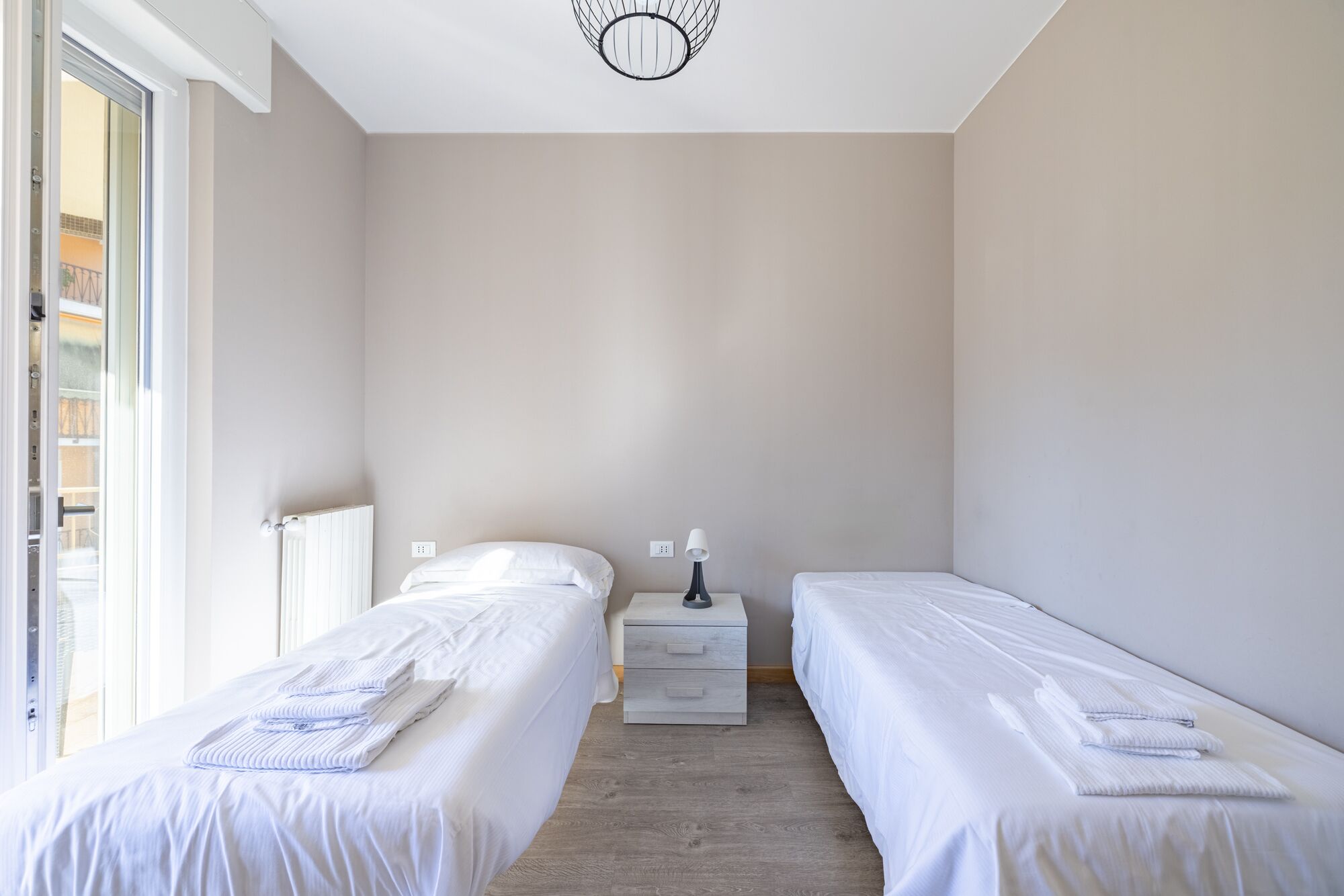 A bedroom with two single beds, a bedside table with a lamp, a radiator, and a window with blinds.