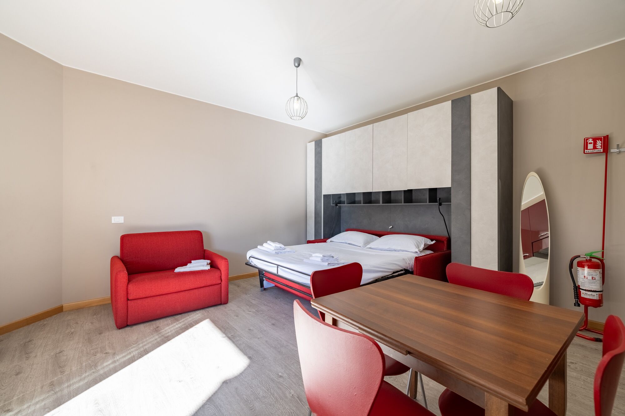 A modern hotel room with a bed, a red sofa, a wooden dining table, and a red chair.