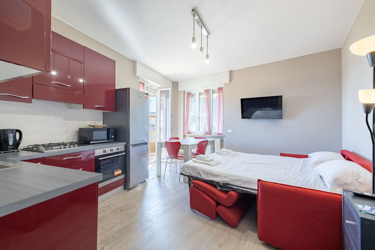 Een compacte hotelkamer met kitchenette, een bed met rode hoofdeinde, een aan de muur bevestigde televisie en een raam met gordijnen.