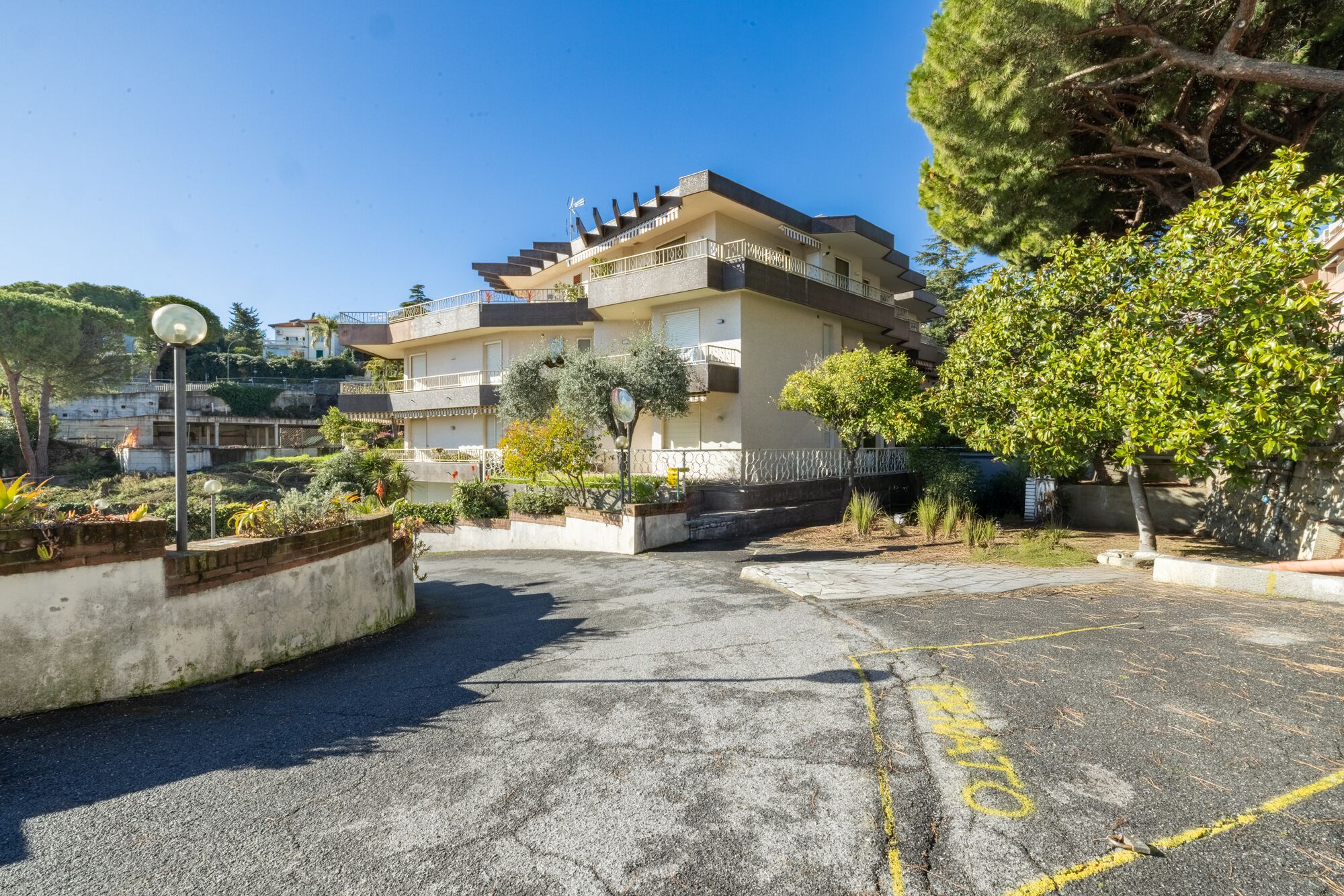 A multi-story residential building with balconies, a parking area with yellow lines, and surrounding greenery.