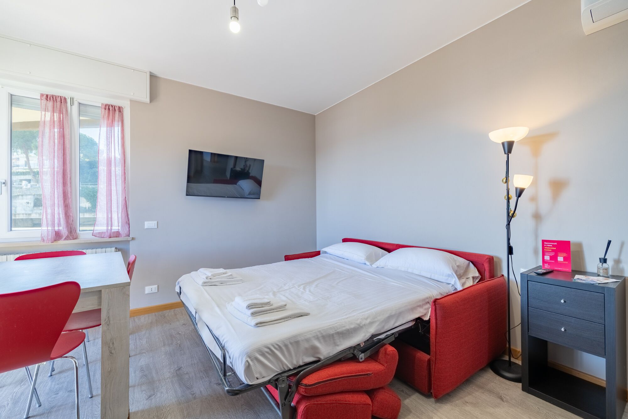 A hotel room with a bed, a desk, a chair, a TV, a lamp, and a bedside table.