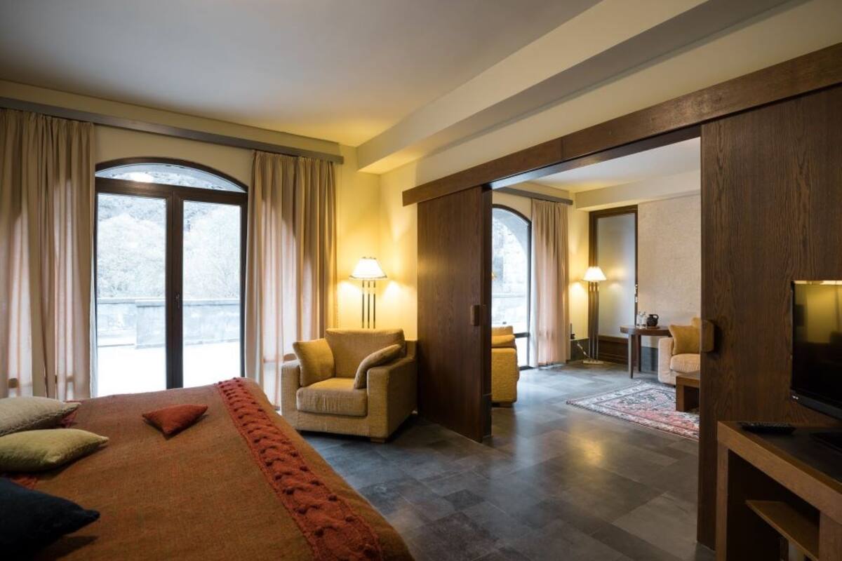 Ein Hotelzimmer mit einem großen Bett, zwei Sesseln, einem Fernseher und Blick ins Freie durch ein großes Fenster.