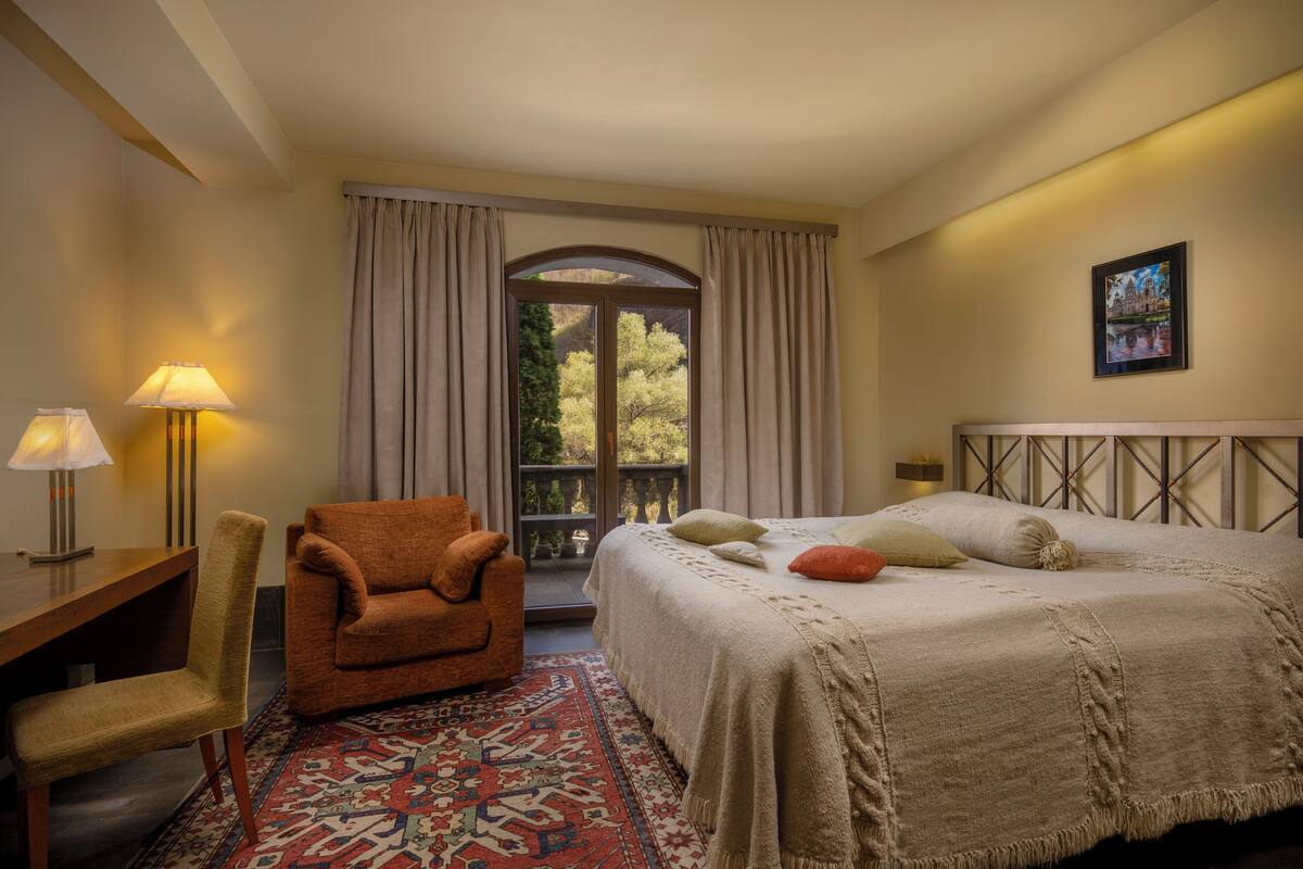 Ein Hotelzimmer mit Bett, Sessel, Schreibtisch, Lampe, einem Fenster mit Vorhängen und einem Balkon.