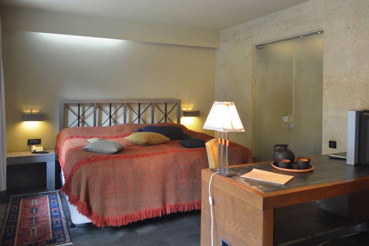 Ein Hotelzimmer mit einem Bett, Nachttischlampen, einem Schreibtisch mit Lampe, einem Spiegel und einem kleinen Tisch mit Wasserkocher und Tassen.