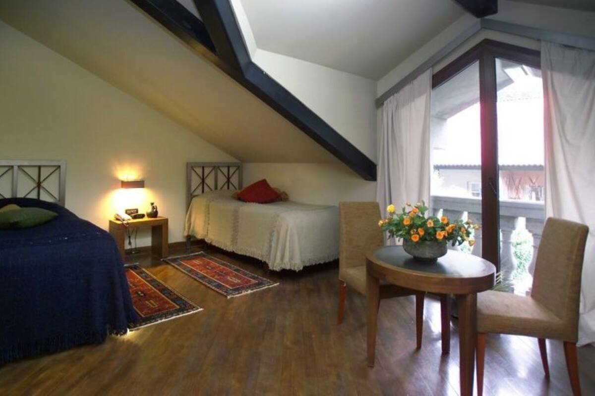 Ein Schlafzimmer mit schräg steilender Decke, einem Bett, einem Nachttisch mit Lampe, einem Sessel und einem kleinen Tisch mit Blumen, sowie ein Blick durch ein Fenster.