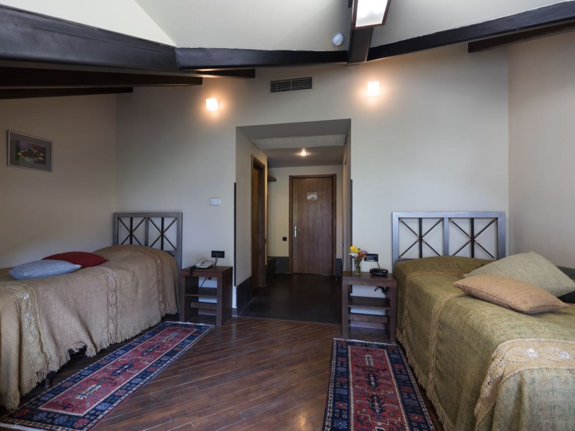 A room with two beds, each with a headboard and a bedside table. There is a door in the center leading to another room.