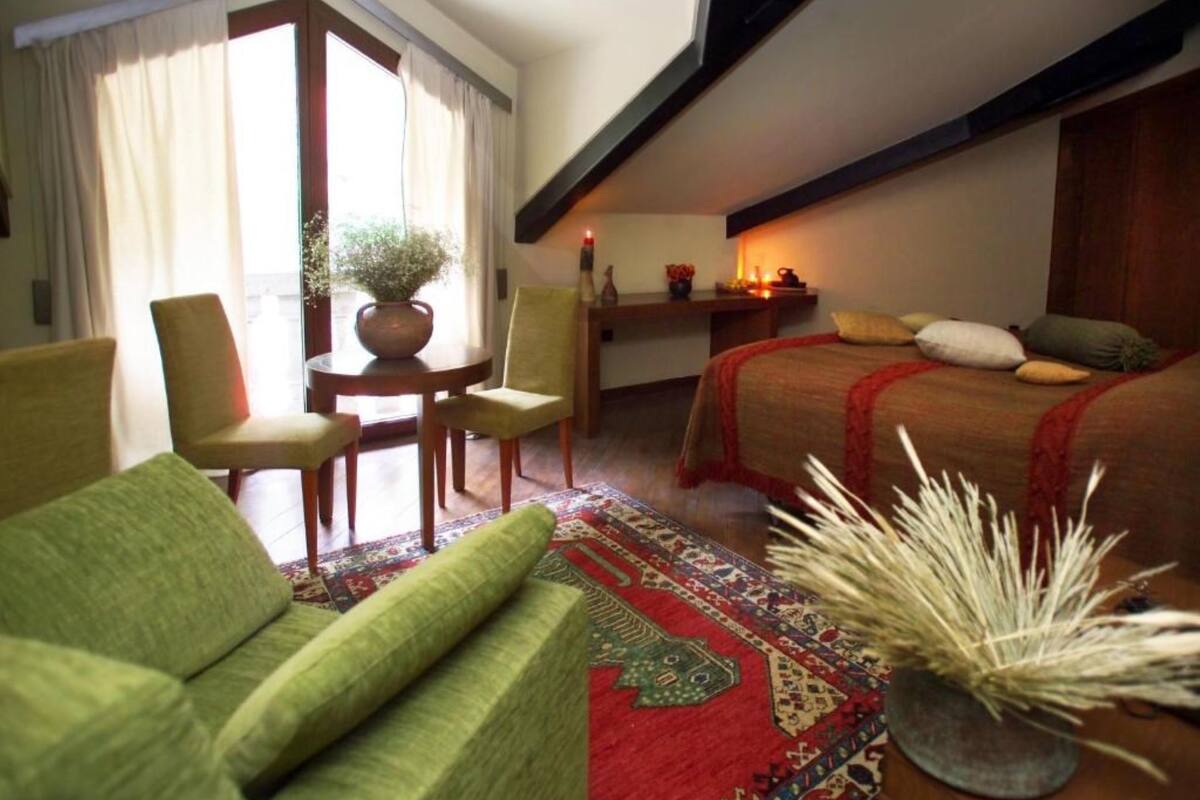 Ein gemütliches Hotelzimmer mit einem grünen Sofa, einem kleinen Esstisch mit Stühlen, einem Bett mit roter Decke und einer Zierpflanze.