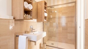Een moderne badkamer met een douchecabine van glas, een witte wastafel en houten planken met handdoeken en toiletartikelen.