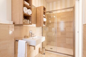 Un baño moderno con ducha de vidrio, lavabo blanco y repisas de madera con toallas y artículos de aseo.