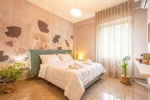 A modern bedroom with a large bed, a bedside table, a potted plant, and a wall with a leaf pattern. - Papavero - Romantic Poppy (Bari)