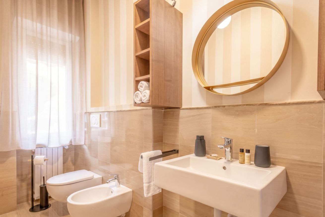 Un bagno moderno con water, bidet e lavandino. Si nota uno scaffale per asciugamani, uno specchio e uno ripiano con asciugamani.