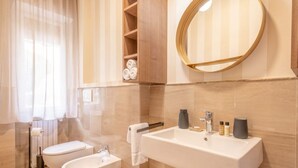Een moderne badkamer met toilet, bidet en wastafel. Er is een handdoekhouder, een spiegel en een plank met handdoeken.