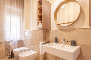 Un baño moderno con inodoro, bidé y lavamanos. Hay un perchero, un espejo y un estante con toallas.