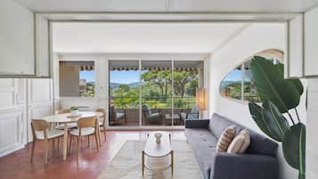 A spacious living area with a sofa, coffee table, and dining set. A large window offers a view of greenery and a distant landscape.