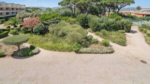 A landscaped garden with various trees and shrubs, a gravel path, and a tennis court in the background. - Apt 2BR/4P-Balcony Saint-Tropez (Saint-Tropez)