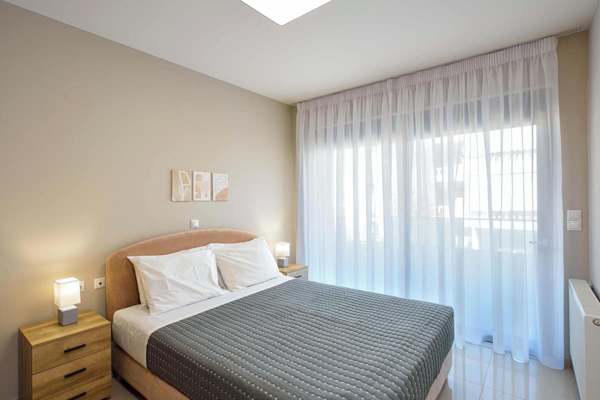 A bedroom with a bed, bedside tables, a nightstand, and a window with sheer curtains.