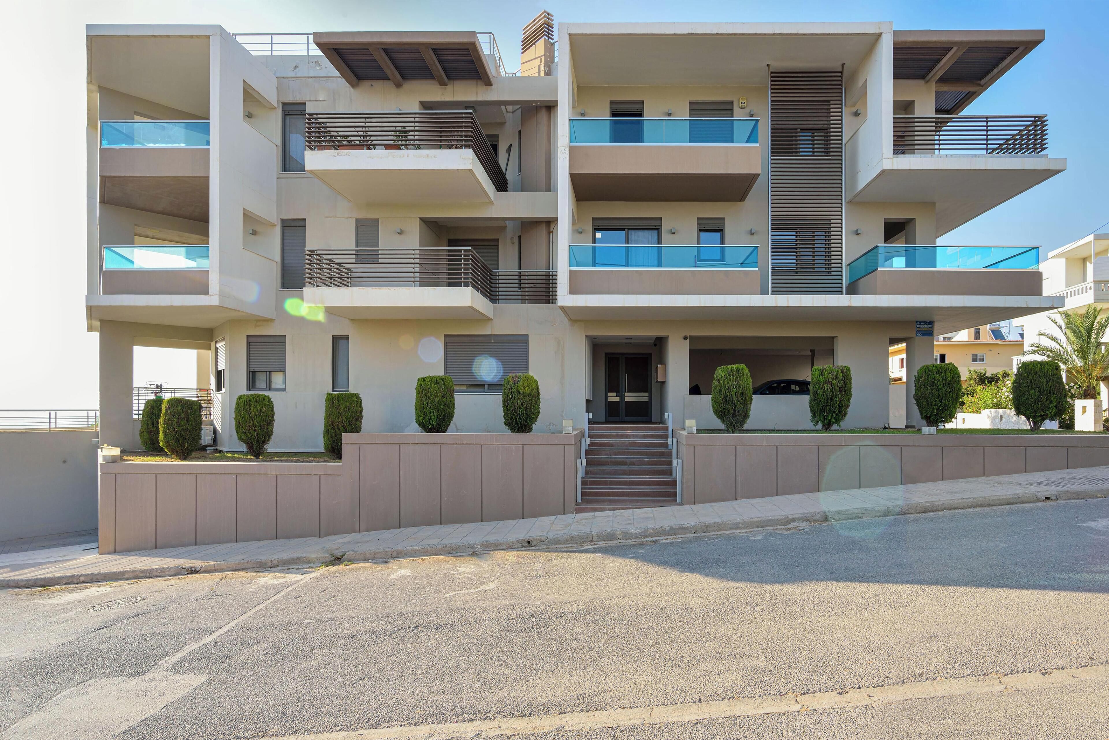 A multi-story residential building with balconies, a central entrance, and a parking area in front.