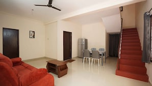 A living room with a red sofa, a wooden coffee table, and a dining area with a table and chairs. There is a refrigerator, a ceiling fan, and a staircase with red steps. - Zenova Stays (Chennai)