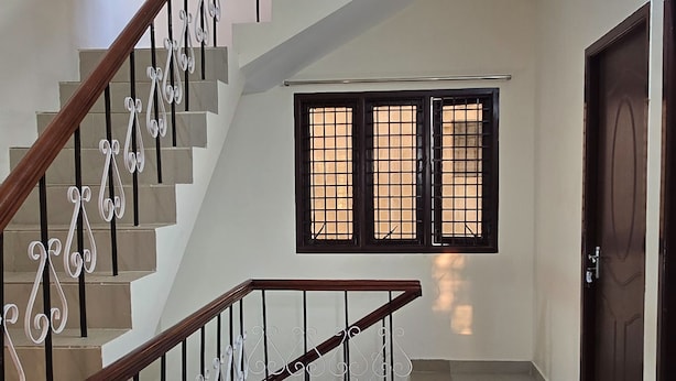 A staircase with a wooden handrail and metal balusters, leading to a landing with a window with barred glass. - Zenova Stays (Chennai)