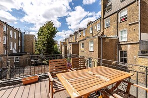 Une terrasse en bois avec une table et des chaises, entourée d’immeubles en briques dont on peut voir les fenêtres et les balcons.
