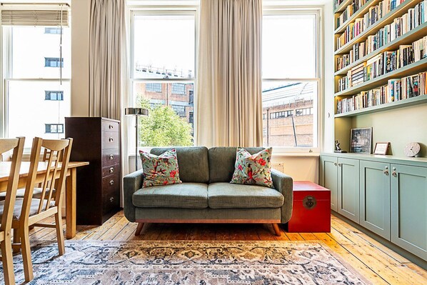 A cozy living room with a green sofa, floral cushions, a wooden dining table, and a bookshelf. - Stylish West Kensington Flat With Character (London)