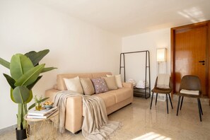 Een woonkamer met een beige bank, een bijzettafeltje met een plant, een klein tafeltje met boeken en een schaal fruit, een zwarte schommelstoel en een houten deur.