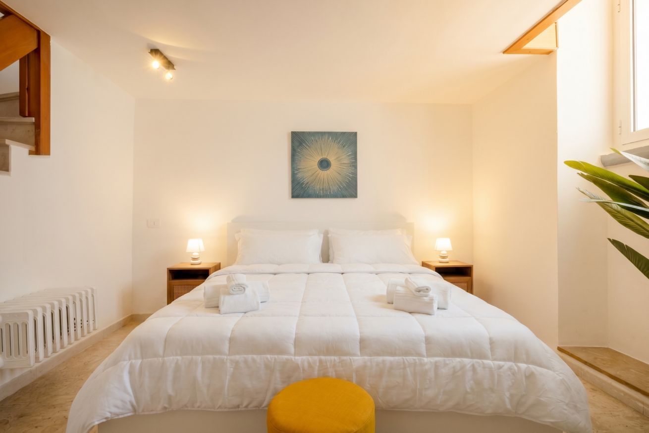 A bedroom with a bed, two bedside lamps, a wall-mounted artwork, a radiator, and a yellow ottoman.