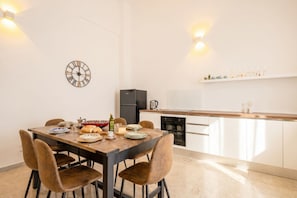 Un coin repas avec une table en bois, des chaises et une horloge murale. Il y a également un coin cuisine avec des placards et un réfrigérateur.
